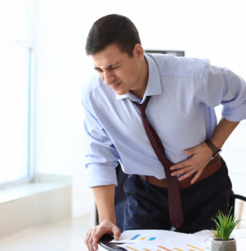 Siedzący tryb życia a problemy ze wzdęciami młody mężczyzna z bólem brzucha stojący przy biurku