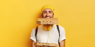 Foodsi – nadwyżka jedzenia nie musi się zmarnować! szczęśliwy mężczyzna na zółtym tle z opakowaniami pizzy jedno opakowanie trzyma w zębach